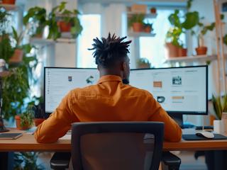 A man sits at a desk, focused on two computer screens displaying various work tasks.