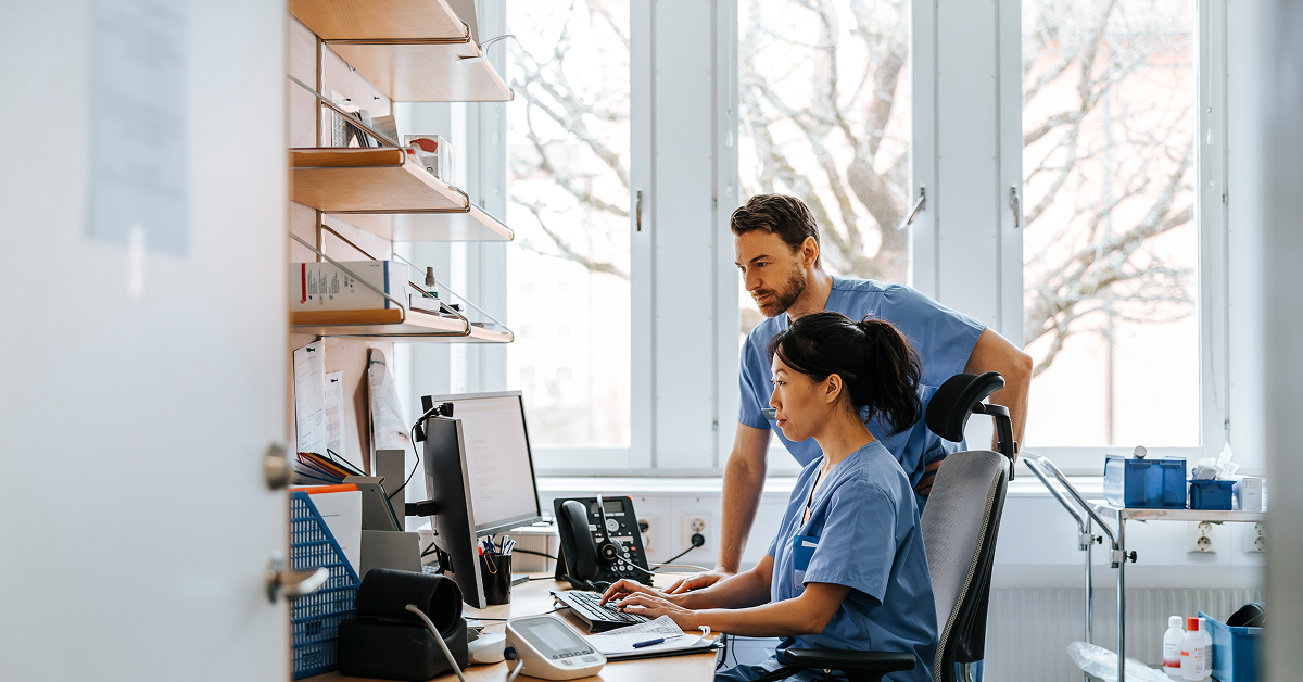 Two healthcare professionals in scrubs reviewing electronic health records on a computer in a clinical office setting.