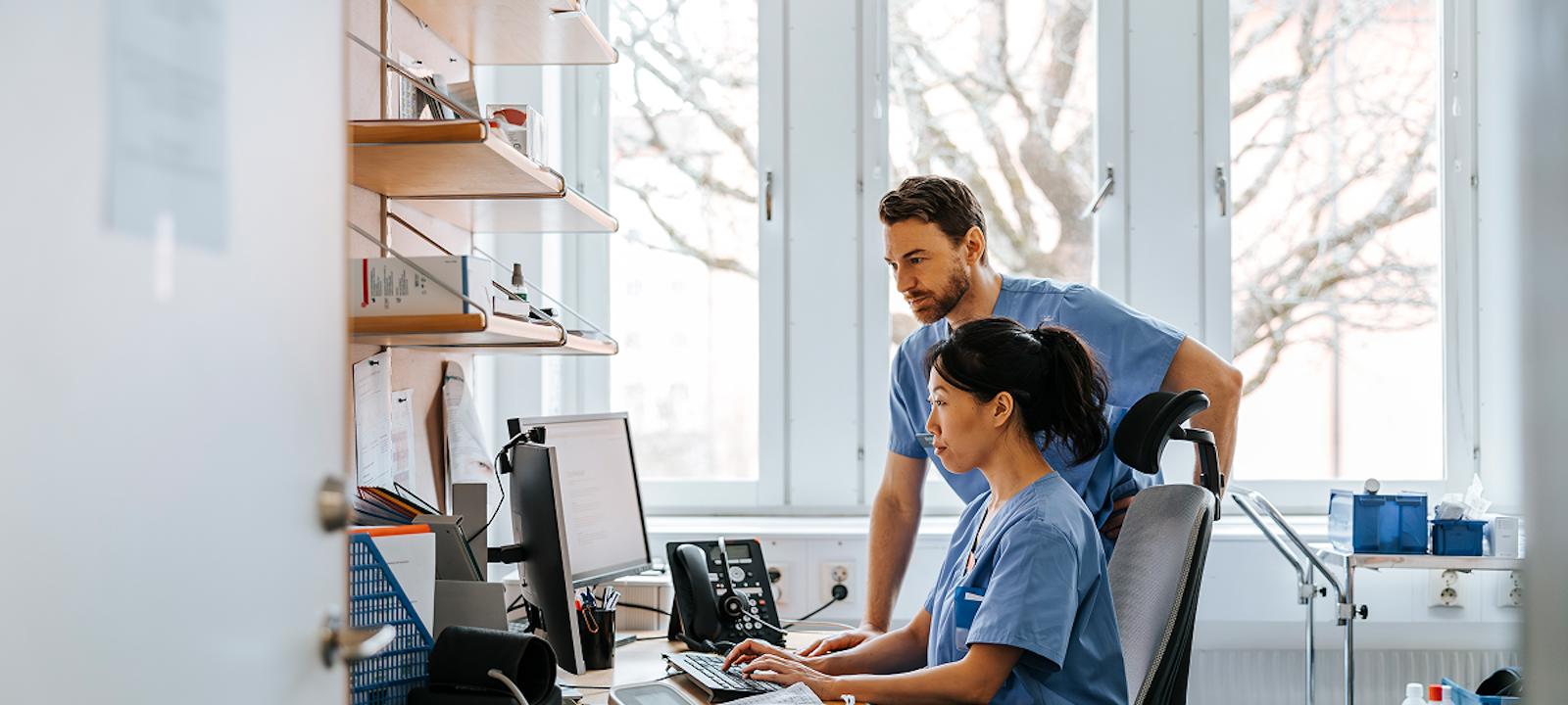Two healthcare professionals in scrubs reviewing electronic health records on a computer in a clinical office setting.