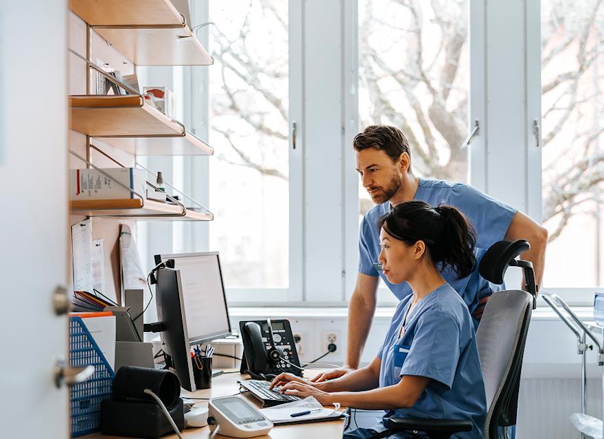 Two healthcare professionals in scrubs reviewing electronic health records on a computer in a clinical office setting.