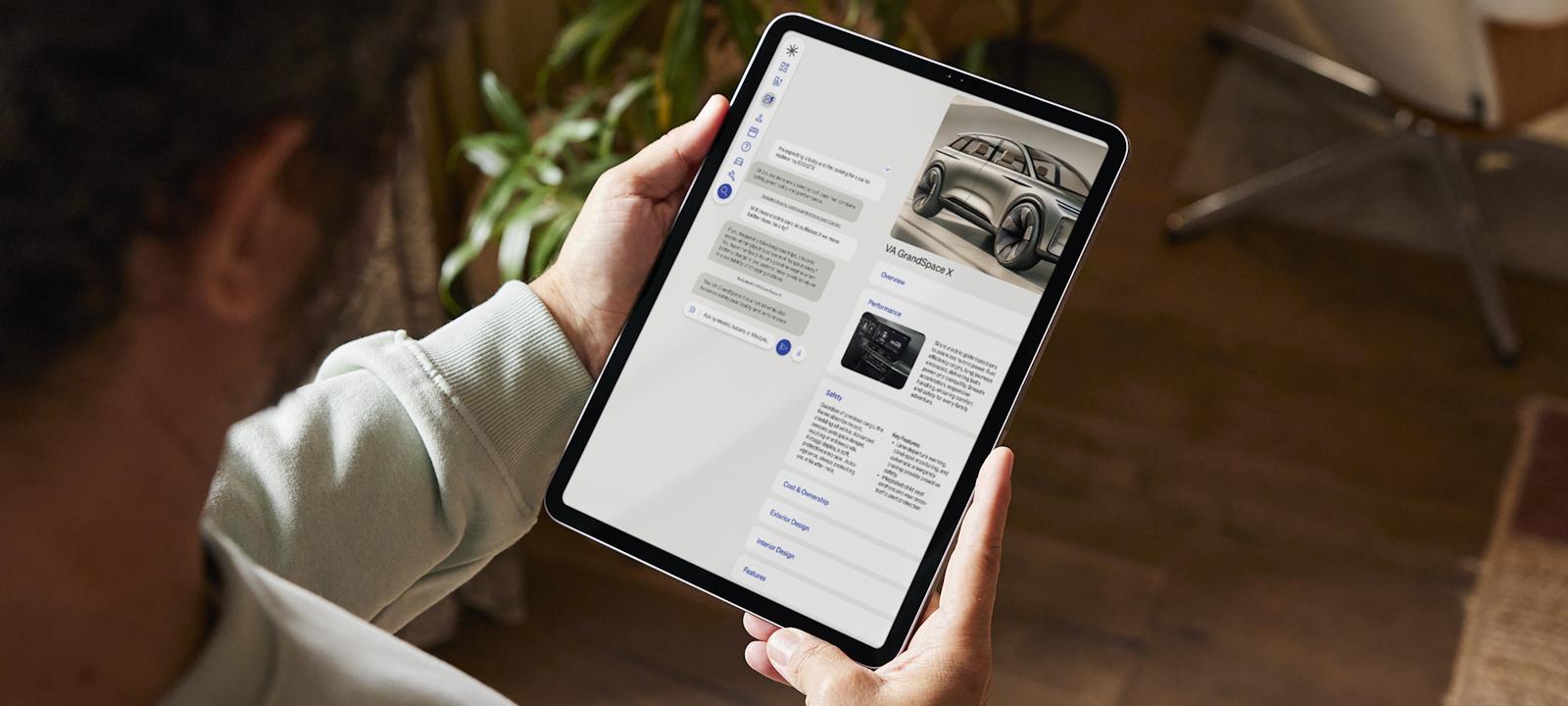 Person holding a tablet while browsing a car information app, displaying details and images of the VA GrandSpace X SUV.