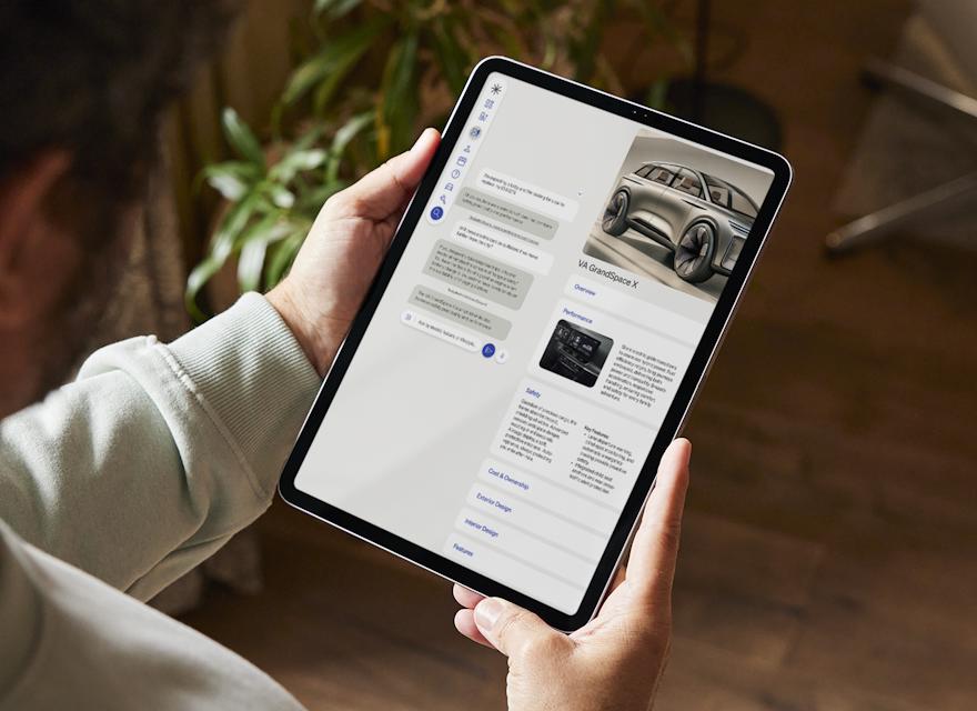 Person holding a tablet while browsing a car information app, displaying details and images of the VA GrandSpace X SUV.