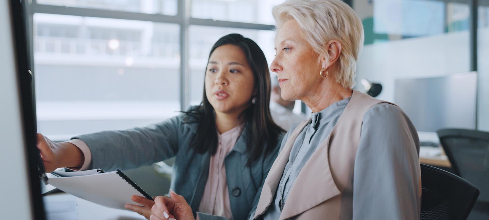 Two professional women reviewing data on a computer screen in a modern office, highlighting teamwork and digital business strategy.