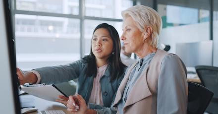 Two professional women reviewing data on a computer screen in a modern office, highlighting teamwork and digital business strategy.