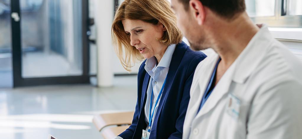A man and woman in a hospital room, both focused on a tablet, discussing its content together
