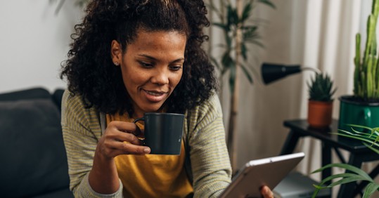 A woman with curly hair smiles while holding a black coffee mug and looking at a tablet screen in a cozy, plant-filled living room