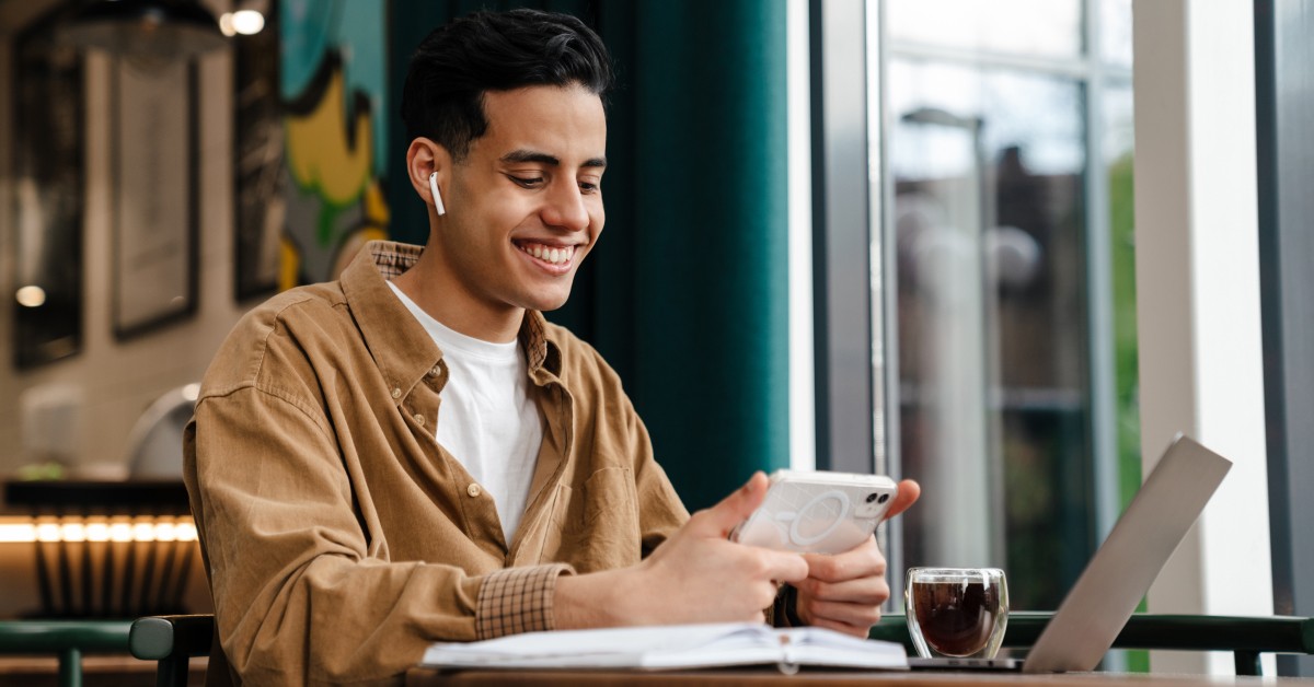 A man with short dark hair, wearing wireless earbuds, is smiling while engaging with his phone at a café table, where a coffee and a notebook are also placed