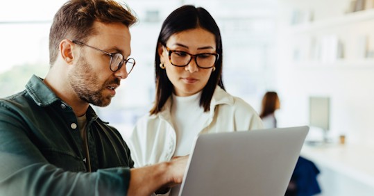 Two colleagues, a man and a woman, both wearing glasses, stand in an office discussing something on a laptop screen, focused on the work at hand