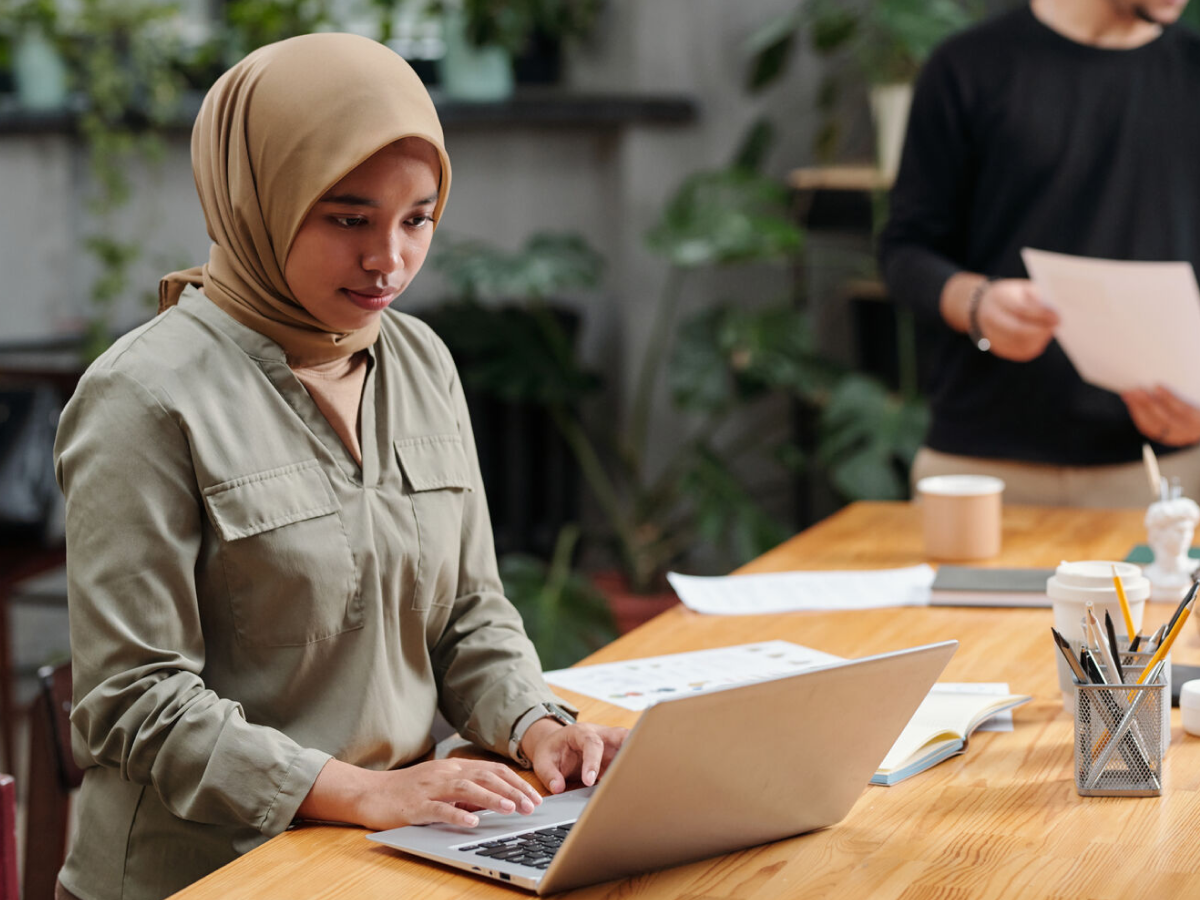 A young woman wearing a beige hijab and olive-green shirt is working on her laptop in a modern office space. In the background, colleagues are holding papers and discussing work at a wooden table filled with office supplies, coffee cups, and documents.