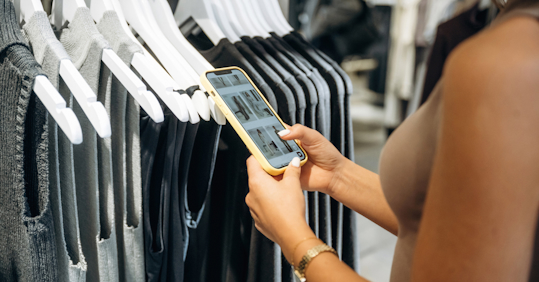 A woman shopping in a clothing store browses an online fashion catalog on her mobile phone while standing in front of a rack of gray and black sleeveless tops. She is wearing a beige sleeveless top and a gold watch, combining in-store shopping with a digital experience.