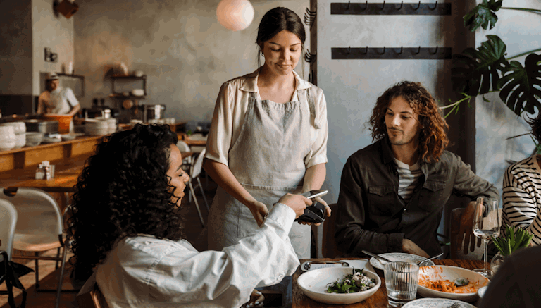 A group of diners at a restaurant are paying for their meal. One woman, seated at the table, is holding a smartphone and using a contactless payment system held by the server, who is wearing an apron. Another person at the table looks on, smiling. The setting is a warm, cozy restaurant with wooden tables and a relaxed atmosphere, where people are enjoying their meal. The background shows additional diners and kitchen staff, contributing to the bustling yet comfortable ambiance.