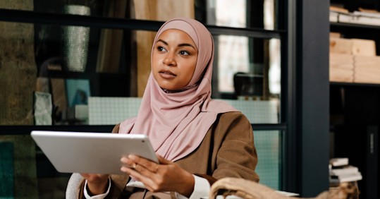 A woman wearing a light pink hijab and a brown jacket is holding a tablet while sitting in a modern indoor setting. She appears thoughtful, gazing into the distance, with shelves and a large window visible in the background. The scene conveys a professional and focused atmosphere.
