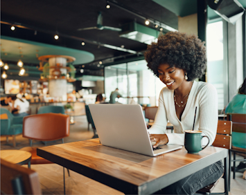 A woman with an afro hairstyle works on her laptop in a modern cafe, smiling with a cup of coffee on the table in front of her