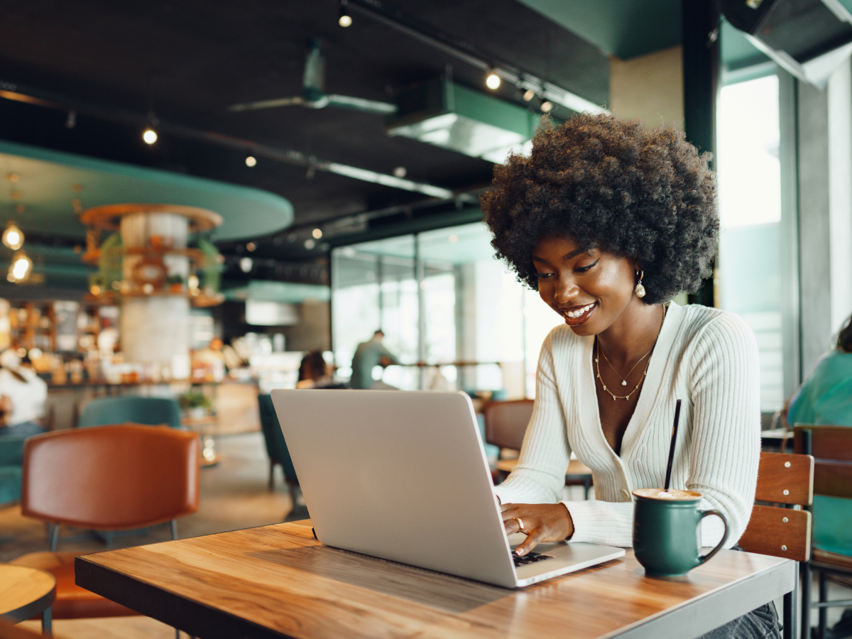 A woman with an afro hairstyle works on her laptop in a modern cafe, smiling with a cup of coffee on the table in front of her