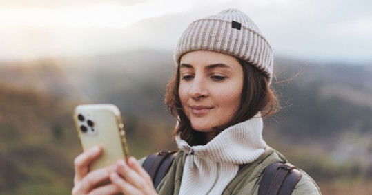 A woman in a knitted beanie and warm jacket, standing outdoors in a scenic area, smiles while looking at her smartphone