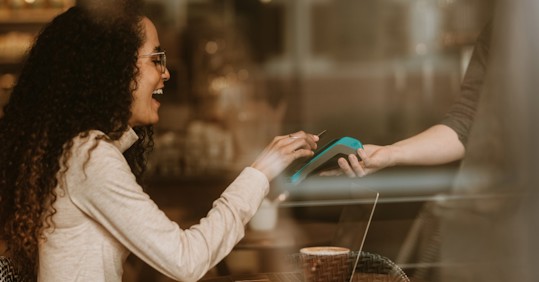 A smiling woman with curly hair holding her phone against a card reader to pay at a cafe, while holding a cup of coffee, with a laptop and notebook on the table