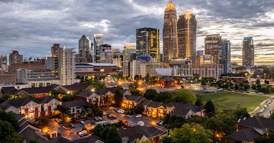 Evening view of Charlotte, North Carolina skyline with illuminated buildings and nearby residential neighborhood, highlighting urban tourism appeal.
