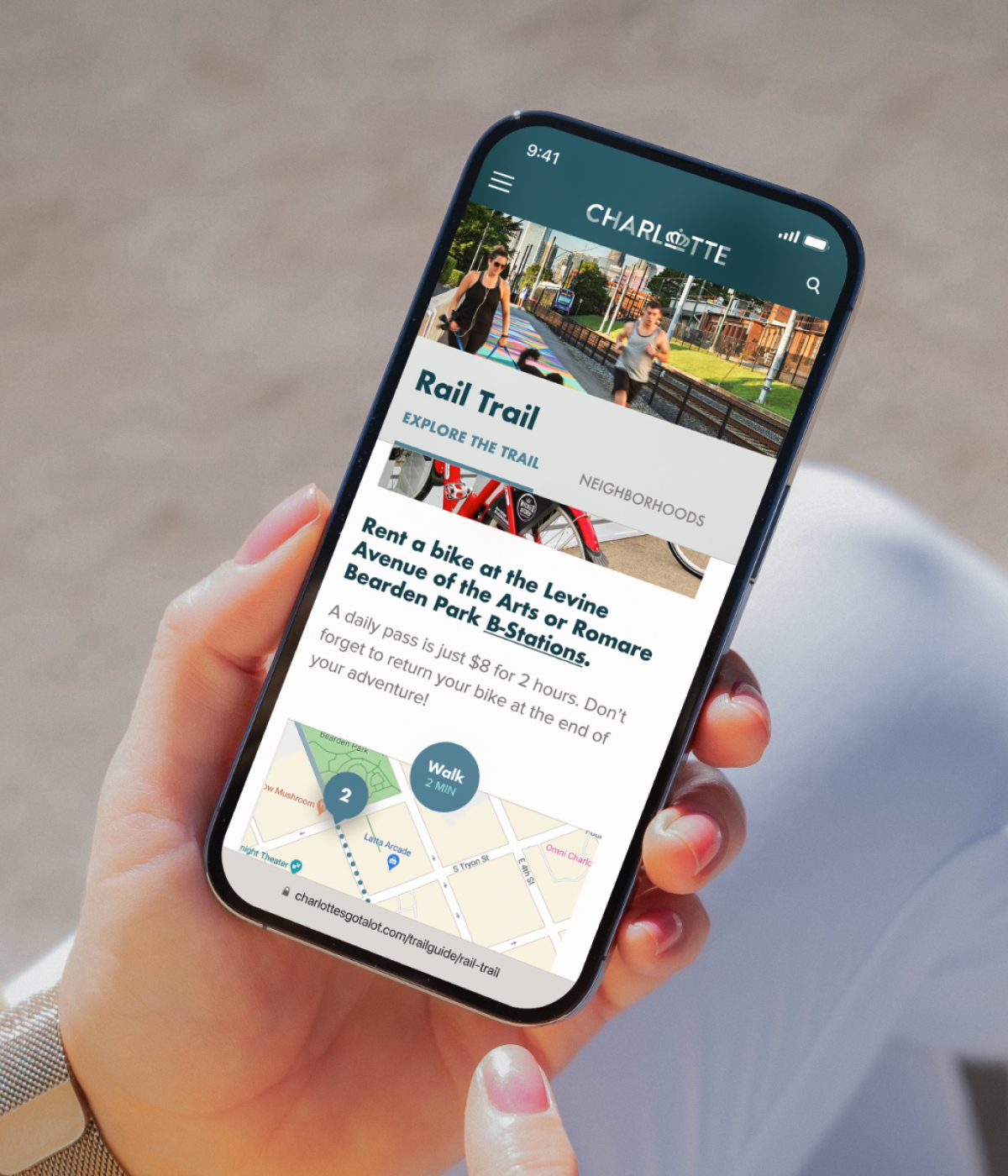Person holding smartphone showing Charlotte’s Rail Trail guide with bike rental info, trail map, and station locations for exploring the city.