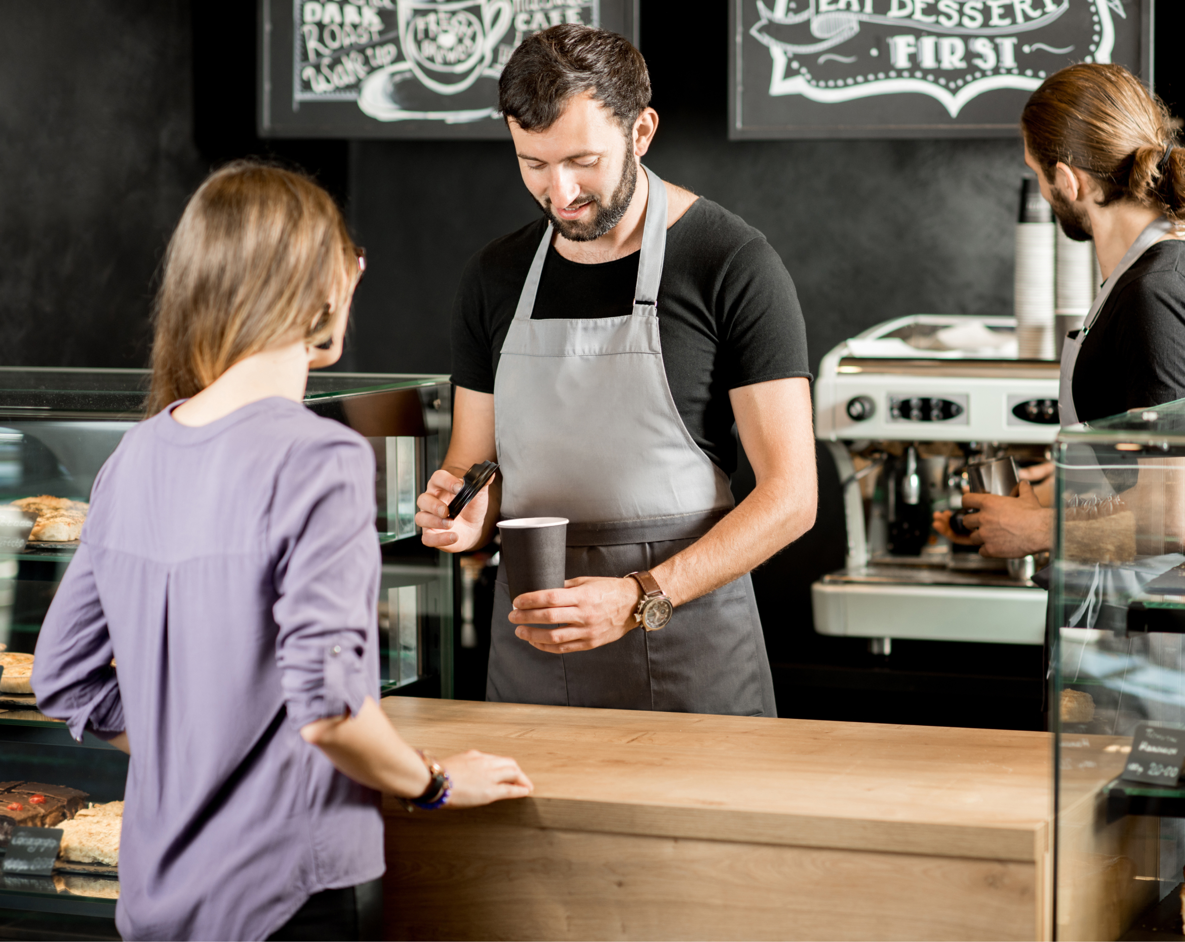 A barista in a gray apron preparing a coffee order for a customer at a modern coffee shop. The customer stands at the counter, while another barista works in the background near an espresso machine. The shop has a cozy, artisanal atmosphere with chalkboard menus in the background.