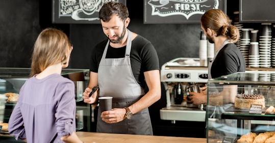A barista in a gray apron preparing a coffee order for a customer at a modern coffee shop. The customer stands at the counter, while another barista works in the background near an espresso machine. The shop has a cozy, artisanal atmosphere with chalkboard menus in the background.
