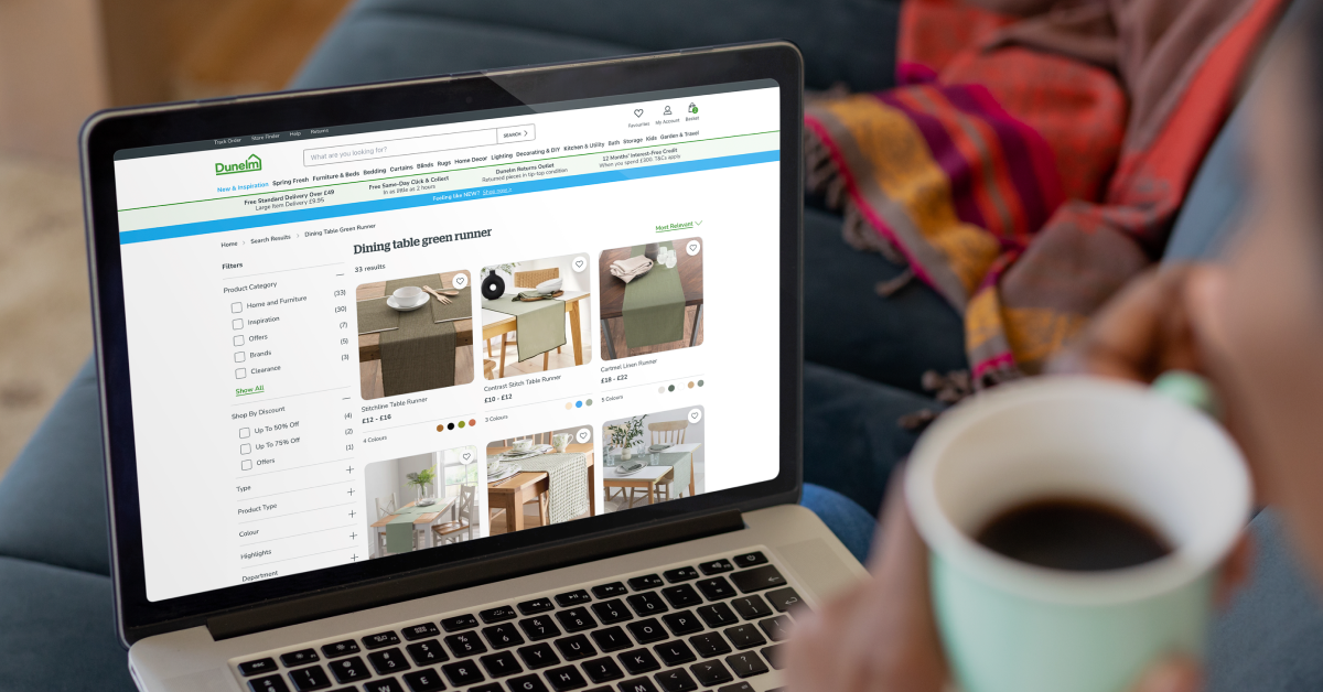 A person holding a cup of coffee while browsing an online store on a laptop. The screen displays a Dunelm website page showing various green dining table runners with product images, prices, and filters for selection. The individual is seated on a couch with a colorful throw blanket in the background.