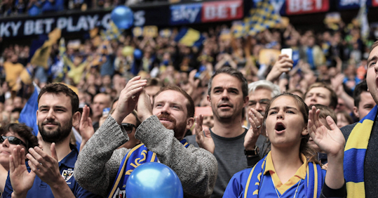 Football fans enthusiastically cheering in the stands, some clapping and others shouting in support, with a sea of yellow and blue colors around them, indicating strong team spirit and unity.