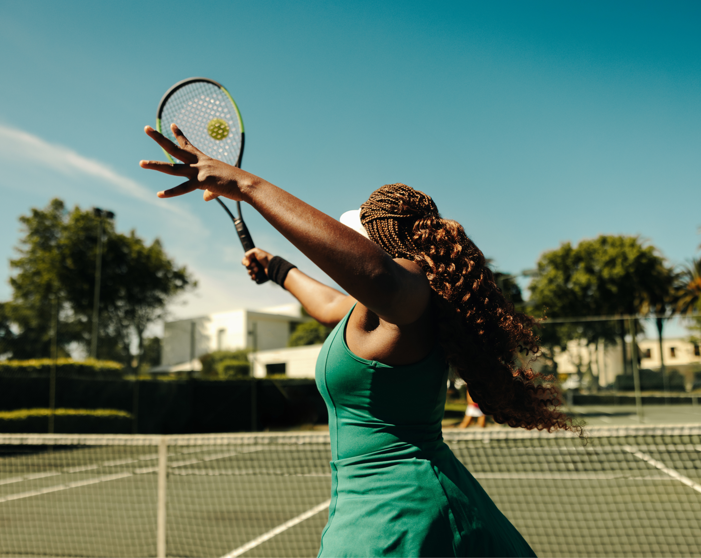 A woman with braided hair is playing tennis on an outdoor court. She is in mid-motion, preparing to serve the ball with her racket. The sun is shining brightly, and the sky is clear, creating a vibrant and energetic atmosphere. The background shows lush greenery and buildings, adding to the picturesque setting. The image captures the athleticism and focus of the tennis player.