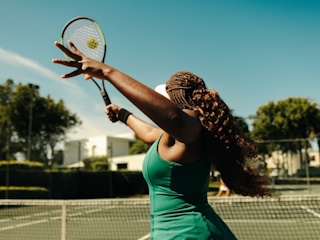 A woman with braided hair is playing tennis on an outdoor court. She is in mid-motion, preparing to serve the ball with her racket. The sun is shining brightly, and the sky is clear, creating a vibrant and energetic atmosphere. The background shows lush greenery and buildings, adding to the picturesque setting. The image captures the athleticism and focus of the tennis player.