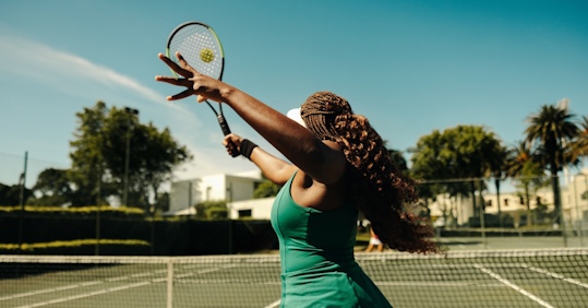 A woman with braided hair is playing tennis on an outdoor court. She is in mid-motion, preparing to serve the ball with her racket. The sun is shining brightly, and the sky is clear, creating a vibrant and energetic atmosphere. The background shows lush greenery and buildings, adding to the picturesque setting. The image captures the athleticism and focus of the tennis player.