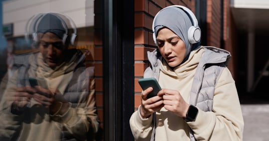 A woman wearing a hijab, headphones, and a puffy vest, standing against a brick wall outdoors, looking at her smartphone while her reflection appears on the glass next to her.