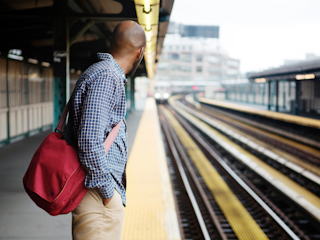 A man wearing a checkered shirt and carrying a red bag stands on a train platform, looking down the tracks as a train approaches in the distance.