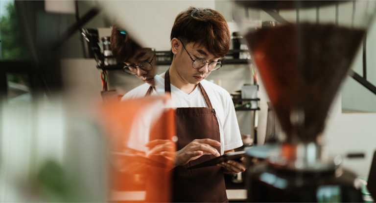 A young chef wearing glasses and a brown apron is focused on a tablet screen in a kitchen. The reflection of the cook is visible in a nearby glass surface, adding depth to the image. The scene suggests the use of technology in the culinary process, perhaps for following recipes or managing kitchen tasks. The kitchen environment appears modern and well-equipped, emphasizing the integration of digital tools in professional cooking.
