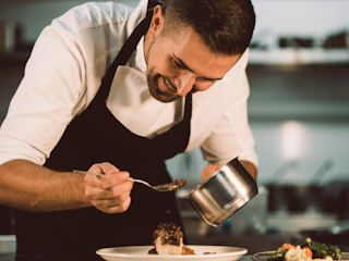 A professional chef is smiling as he carefully plates food in a kitchen. He is wearing a white chef’s coat and a black apron, using a spoon to delicately arrange the dish. The scene captures the chef’s attention to detail and passion for his craft, highlighting the artistry and precision involved in creating a culinary masterpiece. The background suggests a professional kitchen environment, emphasizing the chef’s expertise.