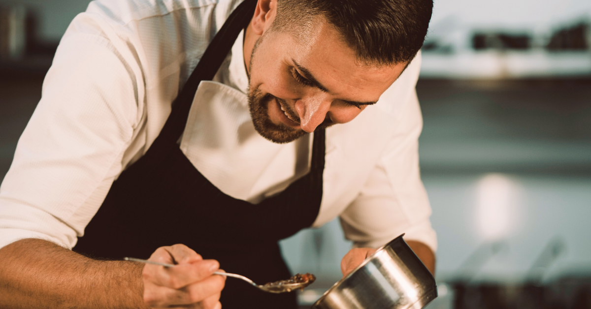 A professional chef is smiling as he carefully plates food in a kitchen. He is wearing a white chef’s coat and a black apron, using a spoon to delicately arrange the dish. The scene captures the chef’s attention to detail and passion for his craft, highlighting the artistry and precision involved in creating a culinary masterpiece. The background suggests a professional kitchen environment, emphasizing the chef’s expertise.