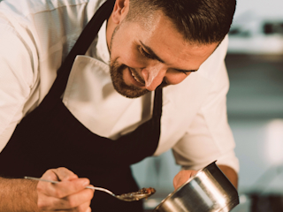 A professional chef is smiling as he carefully plates food in a kitchen. He is wearing a white chef’s coat and a black apron, using a spoon to delicately arrange the dish. The scene captures the chef’s attention to detail and passion for his craft, highlighting the artistry and precision involved in creating a culinary masterpiece. The background suggests a professional kitchen environment, emphasizing the chef’s expertise.