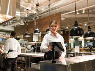 A server in a white shirt standing in a modern, well-lit kitchen, smiling as she interacts with a mounted tablet. The kitchen features industrial-style lighting and stainless steel surfaces, with another staff member working in the background.