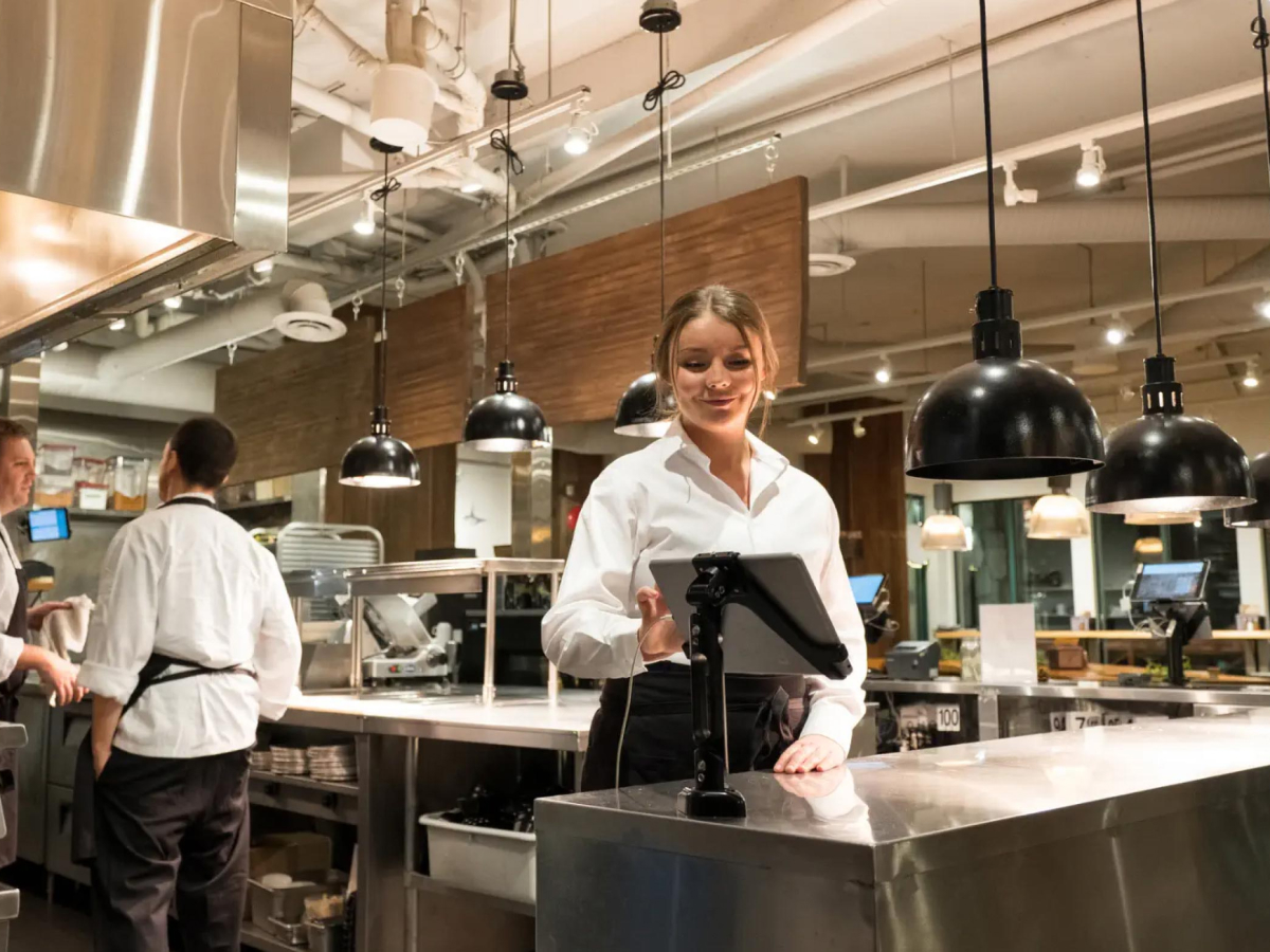 A server in a white shirt standing in a modern, well-lit kitchen, smiling as she interacts with a mounted tablet. The kitchen features industrial-style lighting and stainless steel surfaces, with another staff member working in the background.