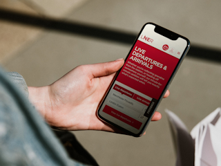 A person holding a smartphone displaying the LNER app, which shows live departures and arrivals information for train stations, with a focus on Manchester Piccadilly and London Euston.