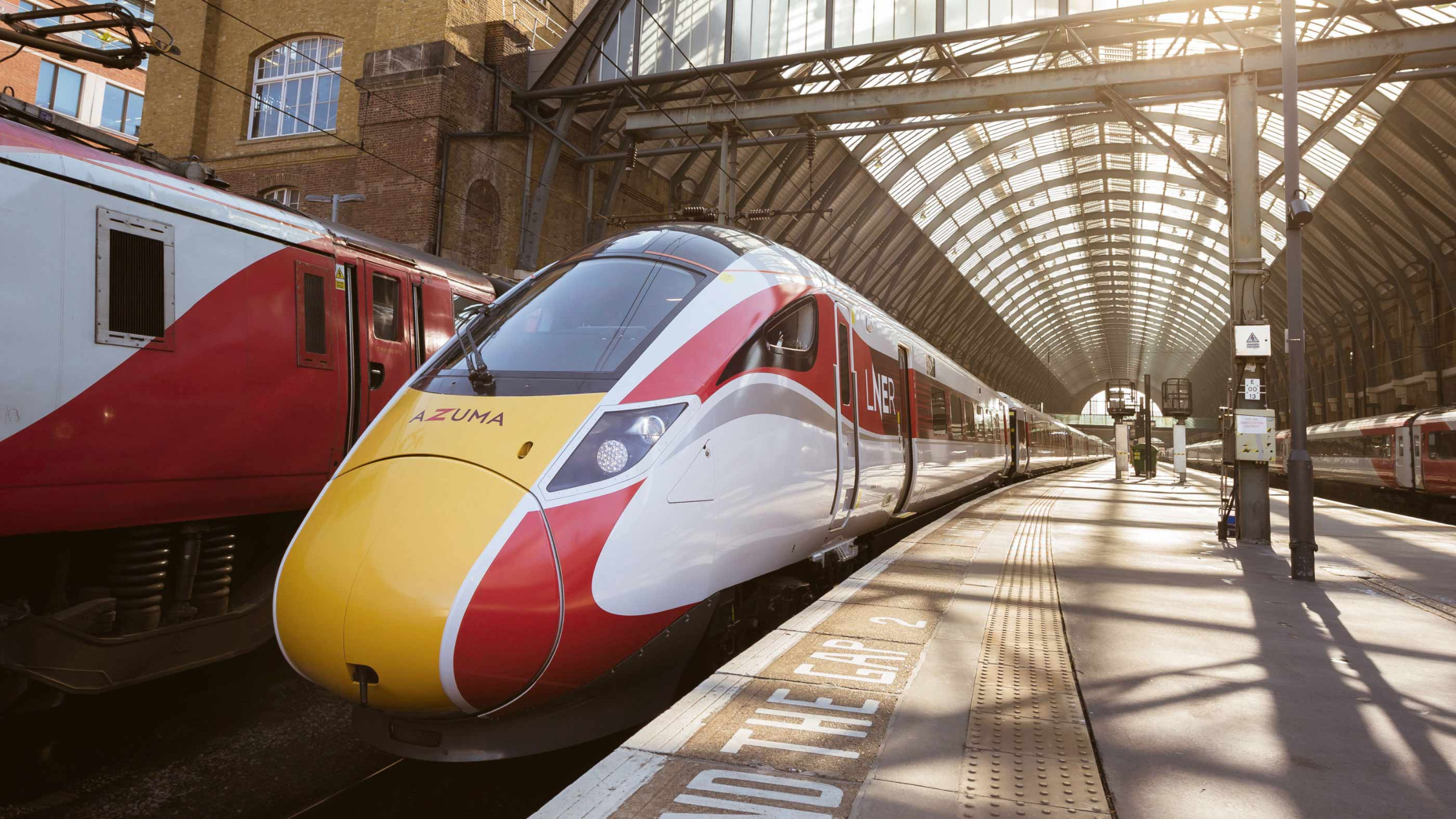 A sleek LNER Azuma train at a sunlit station platform, with the station's arched roof and surrounding architecture visible in the background. The train's modern design and vibrant colors are prominently featured.