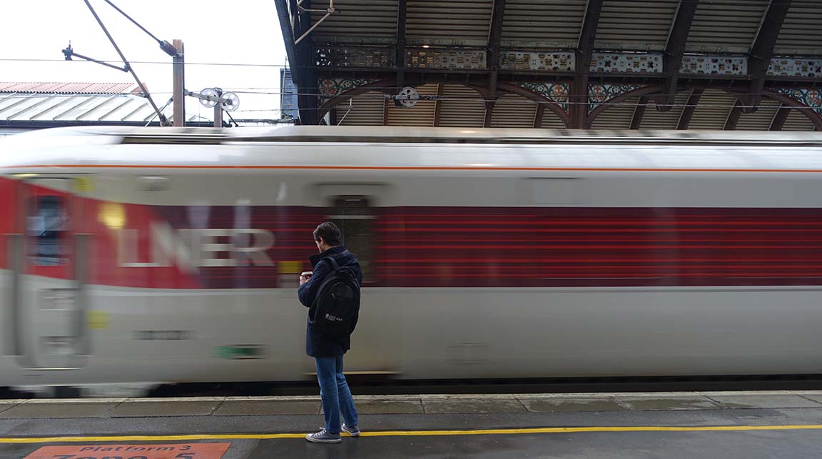 Caucasian man standing in front of a moving silver LNER train which has the LNER logo on a red stripe