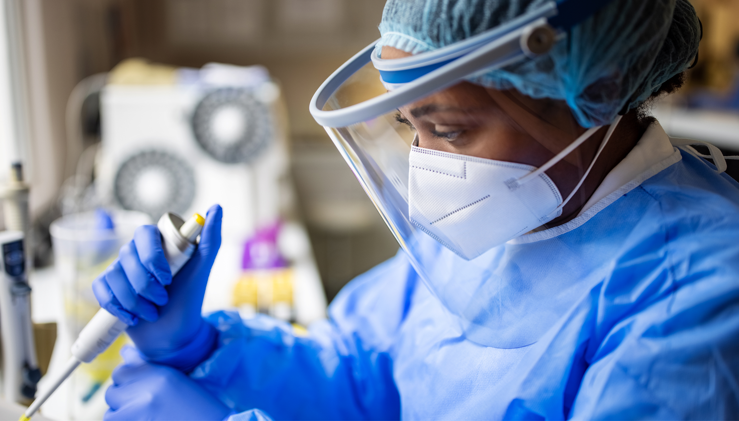 A lab researcher wearing a face shield, surgical cap, mask, gown, and blue gloves carefully holds a pipette over a test tube in a bright laboratory setting, with blurred equipment in the background.