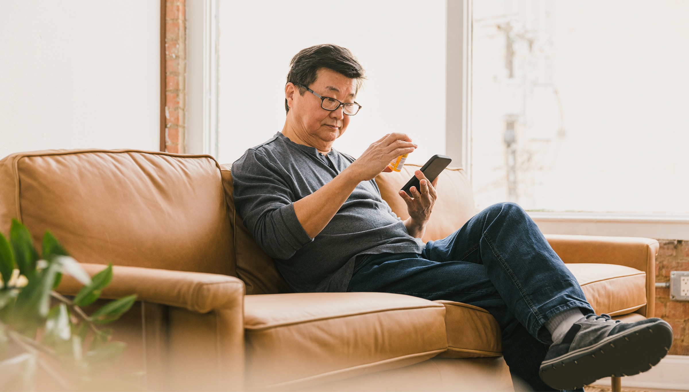 An older man wearing glasses and a gray long-sleeve shirt sits on a tan leather sofa holding a prescription bottle in one hand and his smartphone in the other, reading the label in a bright, modern living room.