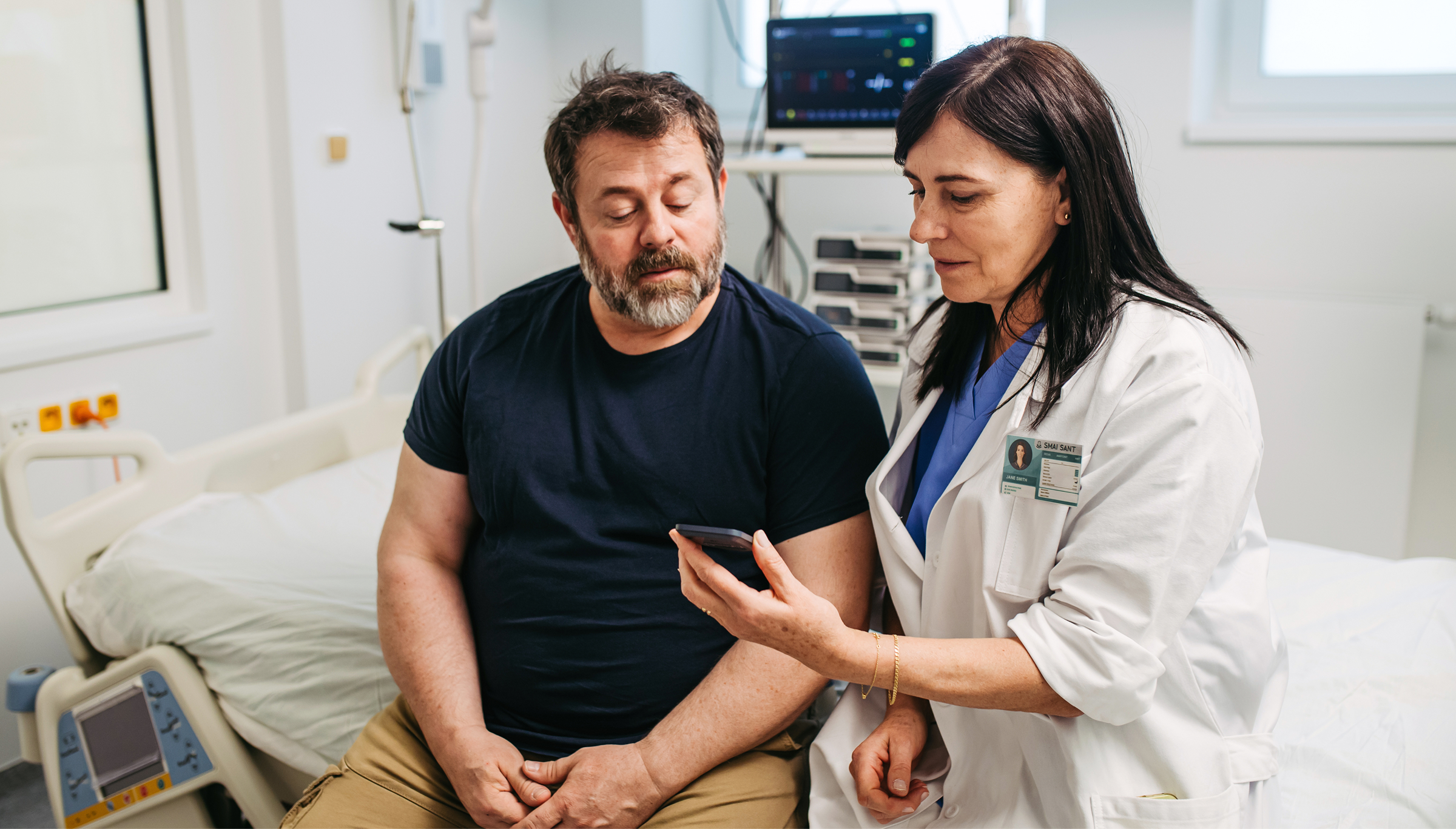A female doctor in a white coat and blue scrubs sits next to a male patient on a hospital bed; she holds up a smartphone showing information while he looks on attentively, with medical monitors and equipment visible in the background.