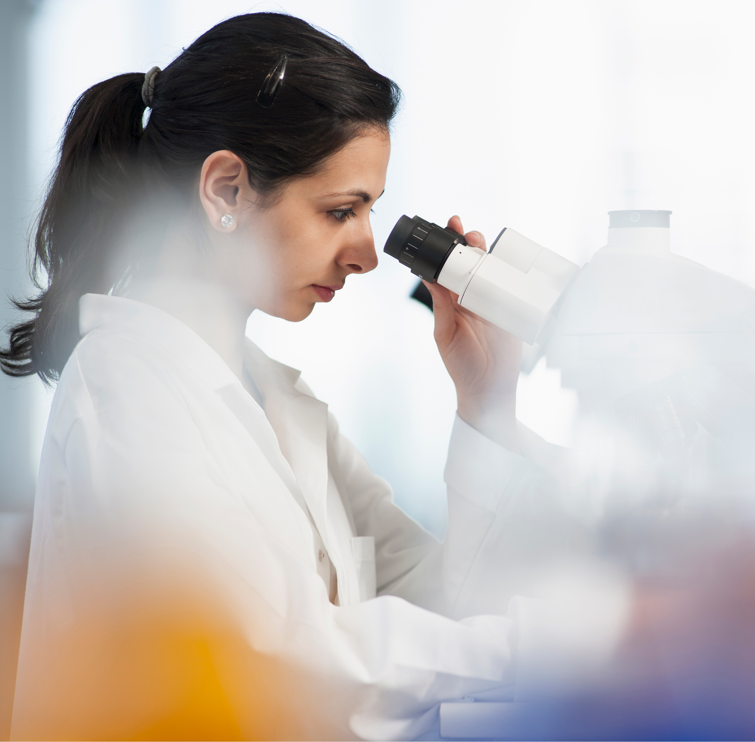 A woman in a white lab coat peers into a white microscope in a bright laboratory, with her dark hair pulled back and her focus on the eyepiece as soft light filters through a window.