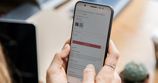 A person holding a smartphone, viewing a product page for Grey Stripe Thinsulate Gloves. The screen shows the product description, price, and purchasing options. In the background, a laptop and a small potted cactus are visible, suggesting a casual or work-from-home environment.