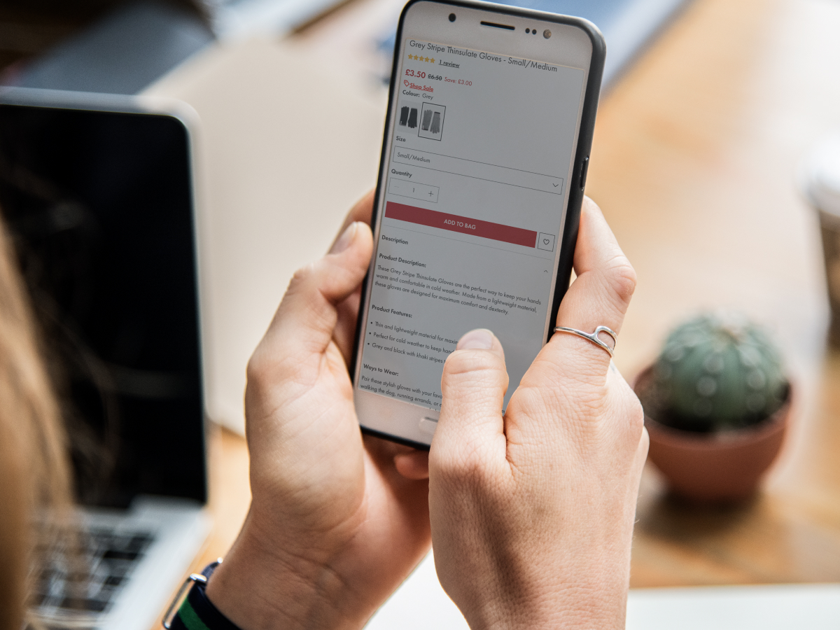 A person holding a smartphone, viewing a product page for Grey Stripe Thinsulate Gloves. The screen shows the product description, price, and purchasing options. In the background, a laptop and a small potted cactus are visible, suggesting a casual or work-from-home environment.