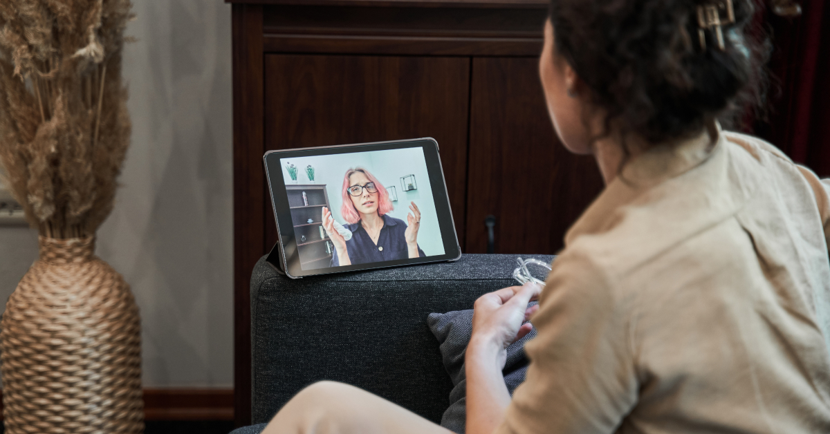 A woman is sitting on a couch, holding her glasses while participating in an online therapy session via a tablet. The tablet screen shows a therapist with pink hair and glasses, speaking and gesturing with her hands. The setting is cozy and intimate, with a vase of dried plants and warm lighting in the background. The scene conveys the convenience and accessibility of mental health support through digital platforms.