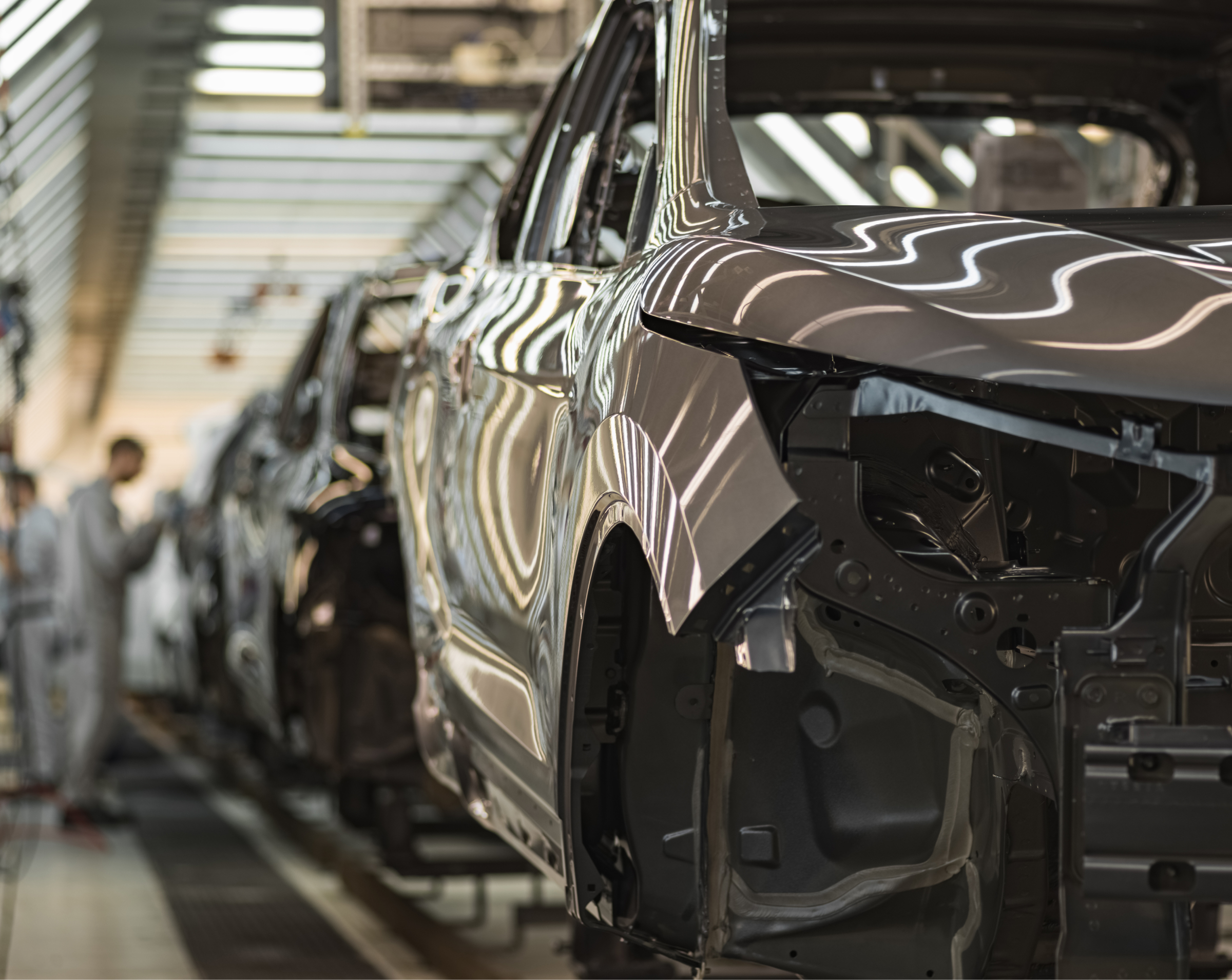 An automotive car assembly line in a factory with several vehicles in various stages of construction and workers blurred in the background.