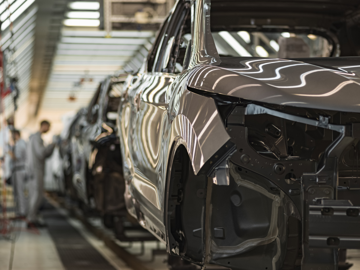 An automotive car assembly line in a factory with several vehicles in various stages of construction and workers blurred in the background.