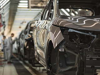 An automotive car assembly line in a factory with several vehicles in various stages of construction and workers blurred in the background.